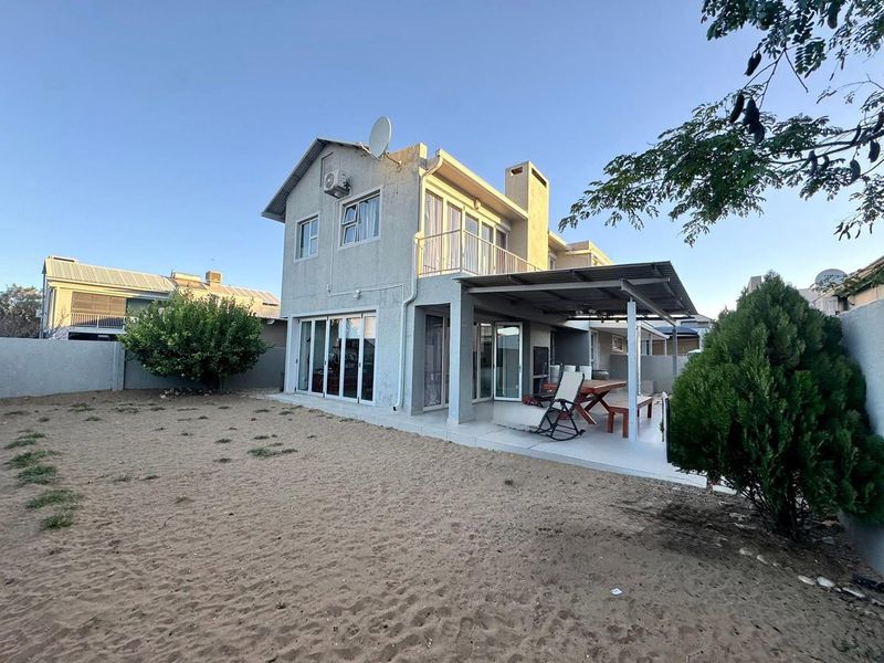 Rear view of a two-story house featuring a covered patio, balcony, and sliding glass doors leading to a sandy backyard area.