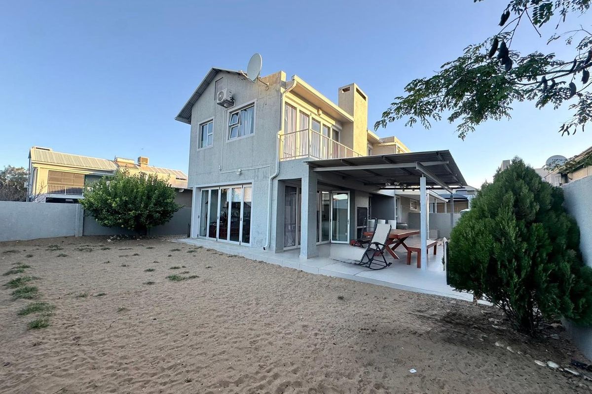 Rear view of a two-story house featuring a covered patio, balcony, and sliding glass doors leading to a sandy backyard area.