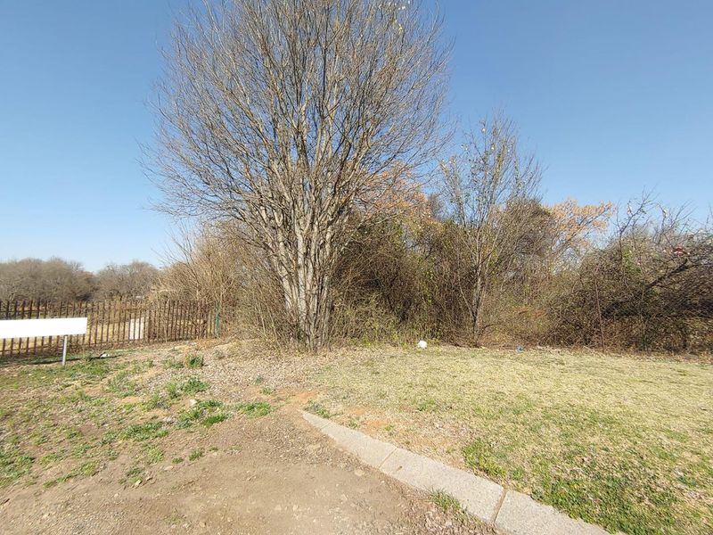 A view of a vacant residential plot featuring mature trees, natural ground cover, and existing metal perimeter fencing.