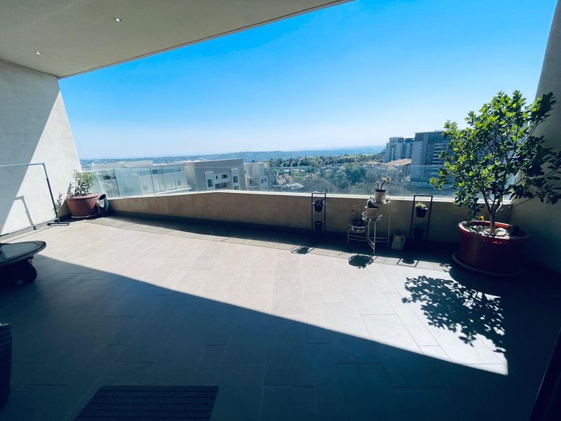 A spacious covered balcony featuring modern tiled flooring, a sleek glass balustrade, and recessed ceiling lighting, offering expansive elevated views.