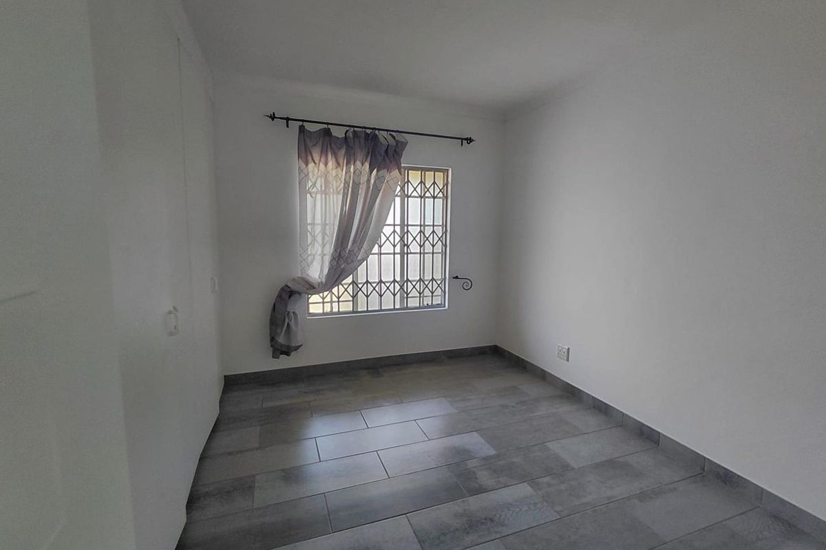 A clean, empty bedroom featuring modern grey tiled flooring, built-in cupboards, and a window fitted with a security gate.
