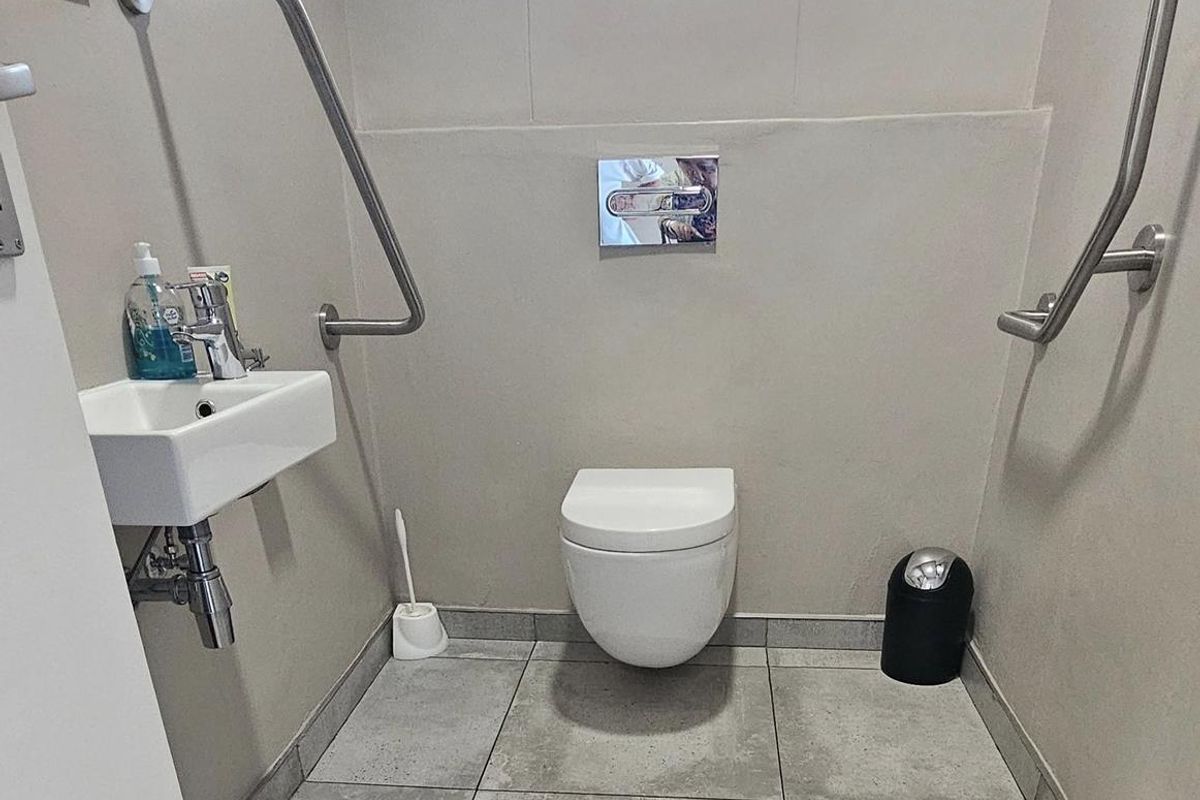 A modern, accessible restroom featuring a wall-mounted toilet, a small wall-hung basin, and stainless steel grab rails for accessibility.