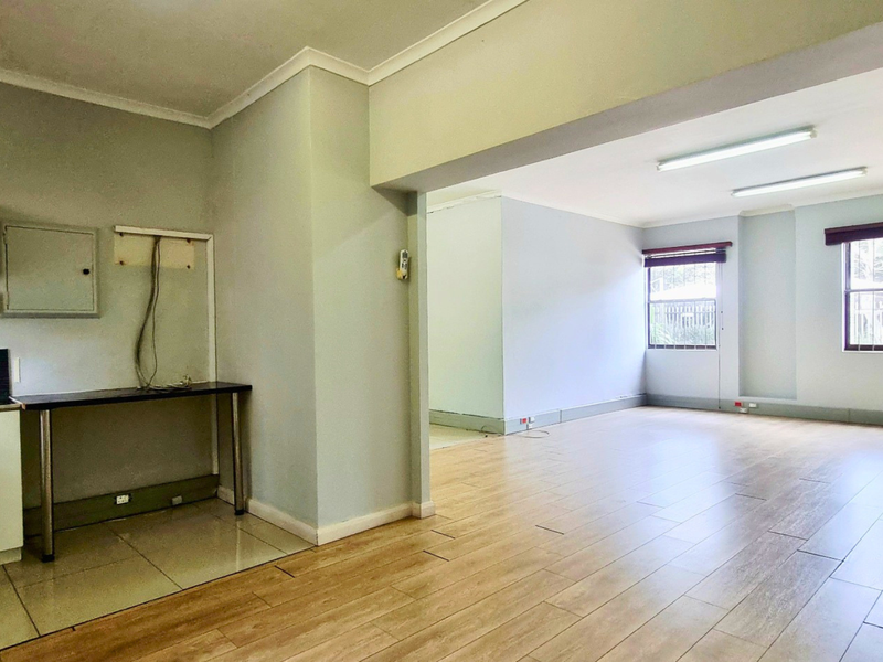 An open-plan office space featuring laminate flooring, built-in kitchenette cabinetry, and large windows with security bars, providing a bright and functional workspace.