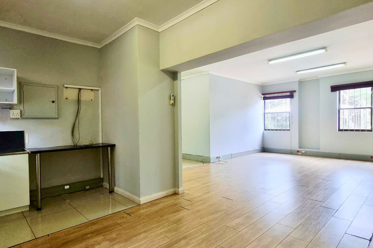 An open-plan office space featuring laminate flooring, built-in kitchenette cabinetry, and large windows with security bars, providing a bright and functional workspace.