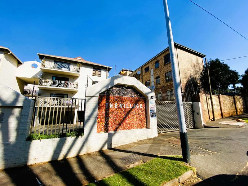 Front exterior view of The Village apartment complex, featuring a secure gated entrance, brick boundary wall, and multi-story residential building with balconies.