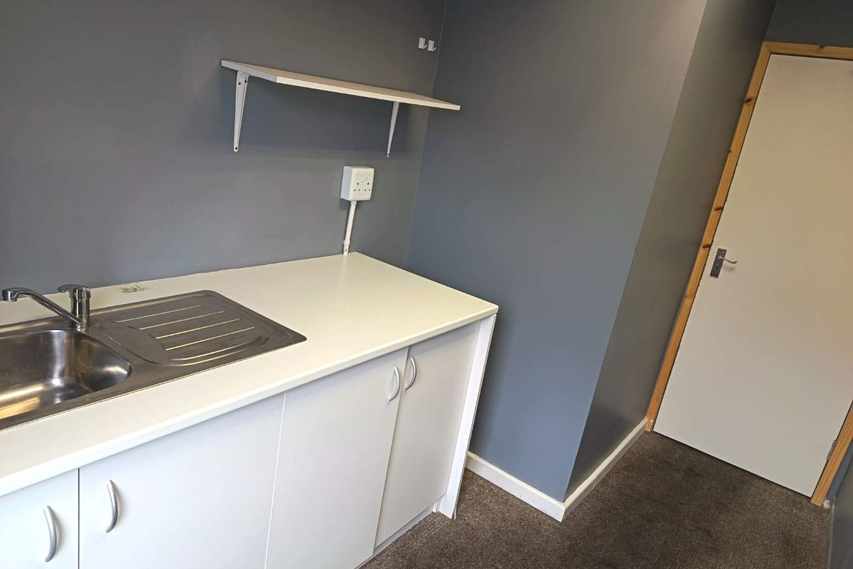 A functional kitchenette area featuring a stainless steel sink, white cabinetry, and a wall-mounted shelf, suitable for office or utility use.