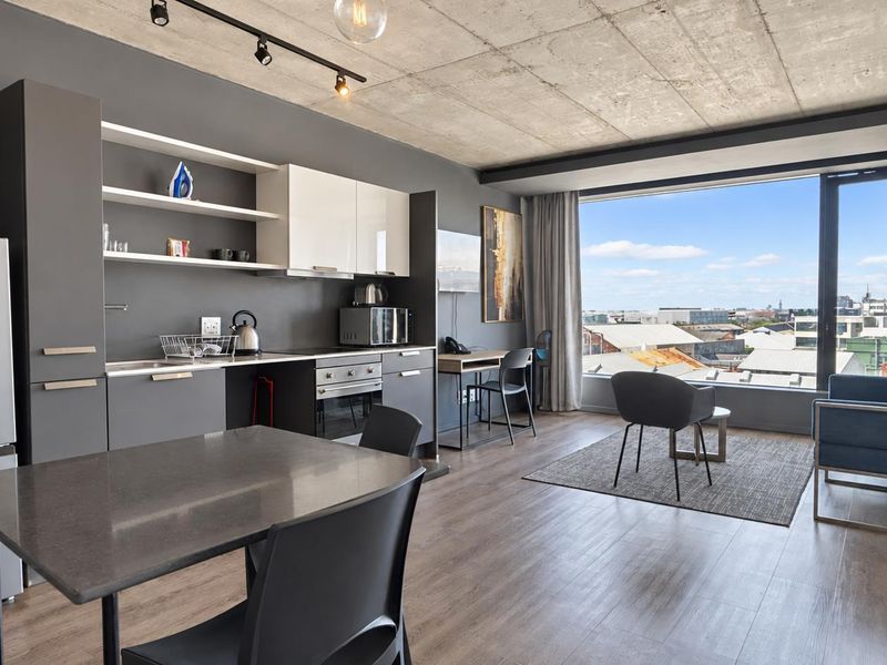 Modern open-plan apartment featuring an exposed concrete ceiling, sleek kitchen cabinetry, and expansive windows offering panoramic urban views.