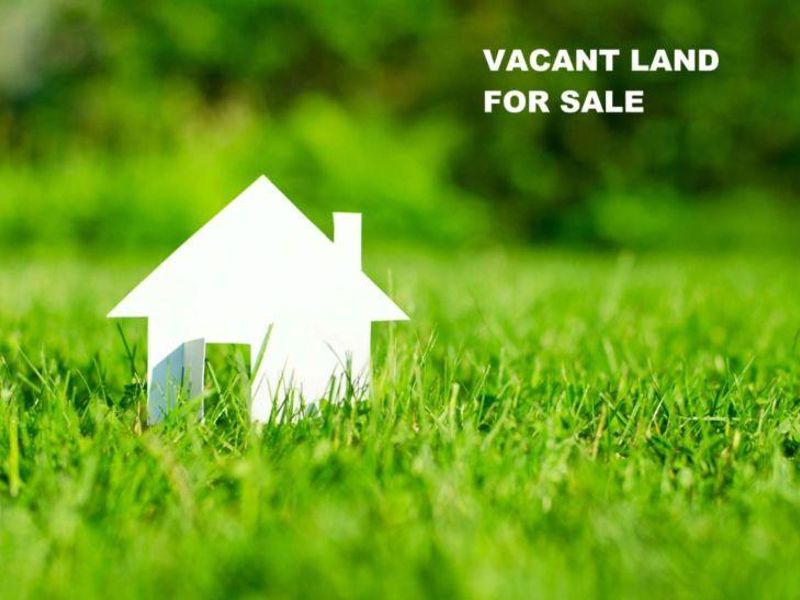 A promotional image for a vacant residential land plot featuring a paper house cutout on a grassy lawn.
