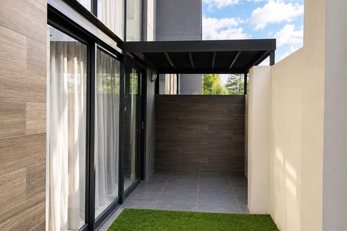 A modern, low-maintenance rear courtyard featuring a covered patio area, tiled flooring, and a small artificial grass lawn adjacent to large glass sliding doors.
