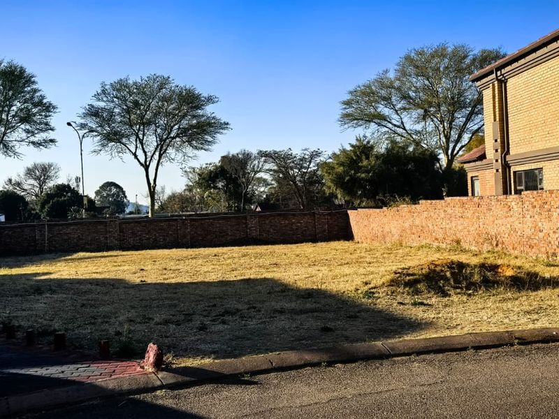 A vacant residential plot featuring a brick boundary wall and paved driveway edge, situated adjacent to an existing brick building.