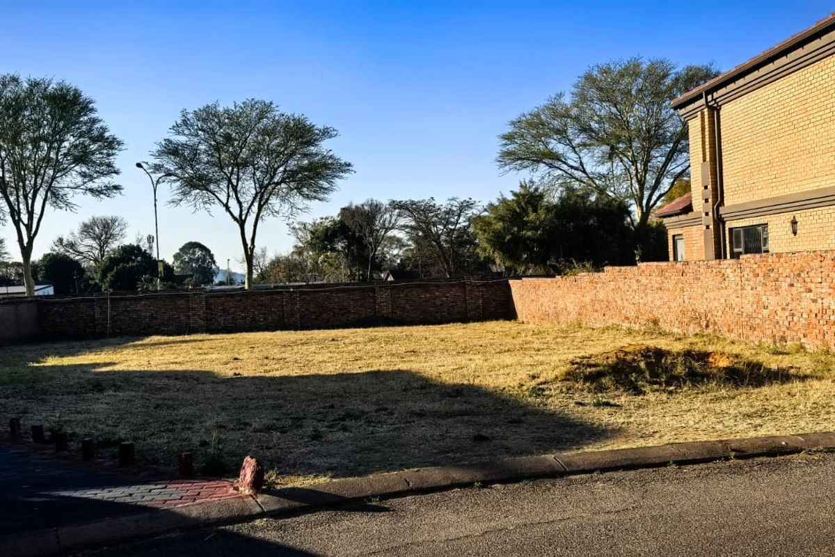 A vacant residential plot featuring a brick boundary wall and paved driveway edge, situated adjacent to an existing brick building.