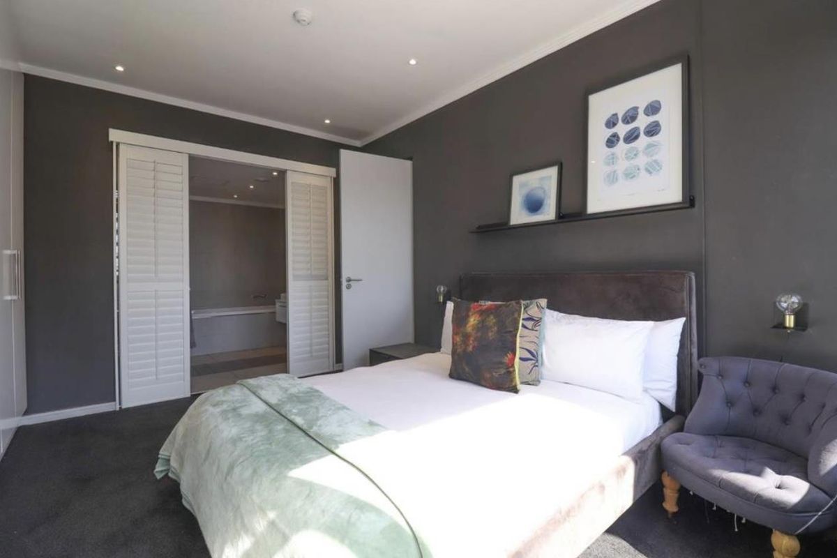 A well-appointed bedroom featuring dark accent walls, recessed lighting, and sliding plantation shutters leading to an en-suite bathroom.