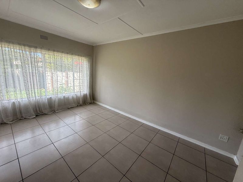 A bright, empty living room featuring neutral wall paint, large windows with natural light, and clean tiled flooring throughout.