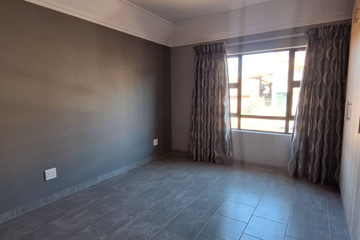 A clean, empty bedroom featuring modern grey tiled flooring, built-in wooden cupboards, and a large window with curtains.