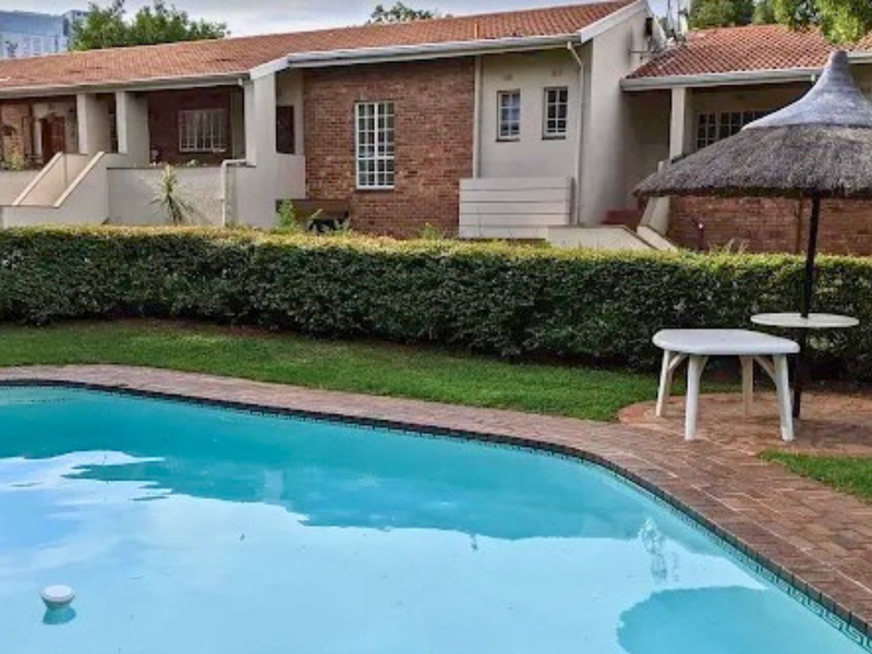 A well-maintained communal swimming pool area featuring brick paving, lush hedged landscaping, and a view of the apartment complex exterior.