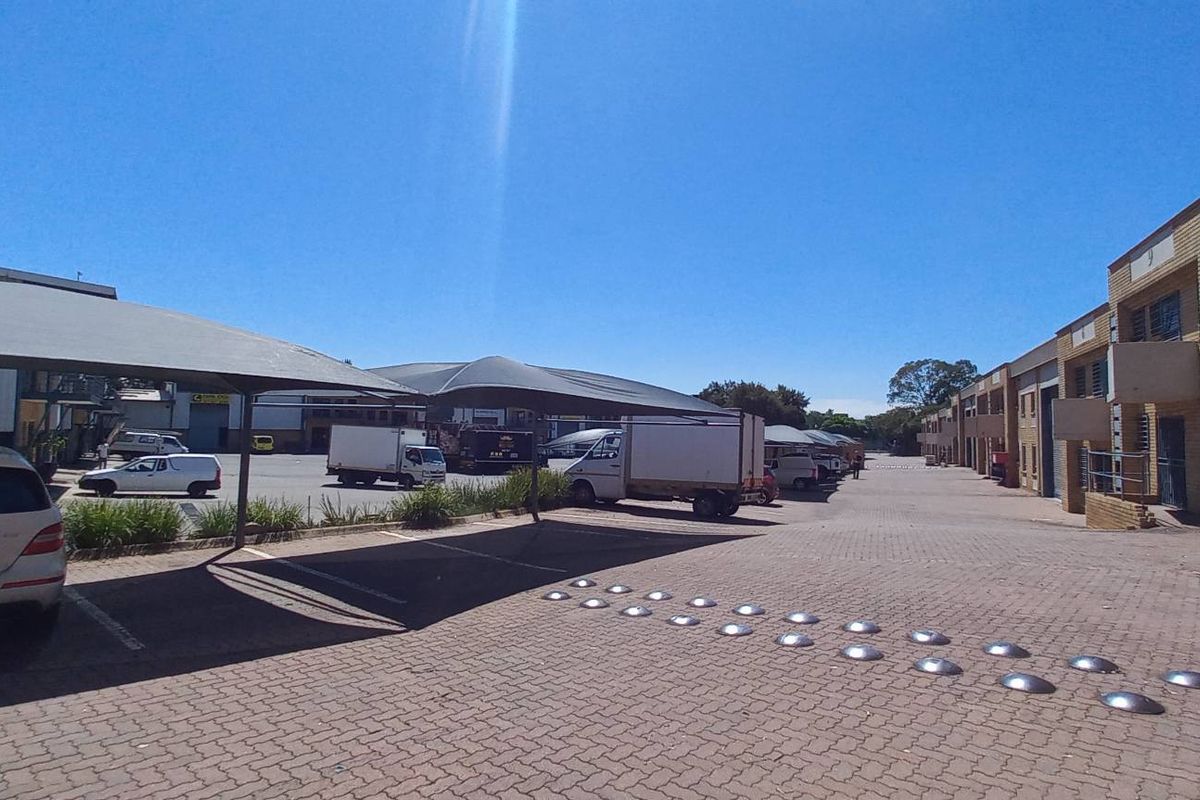 Industrial complex exterior featuring a paved driveway, shade cloth carports, and multiple roller shutter doors for warehouse access.