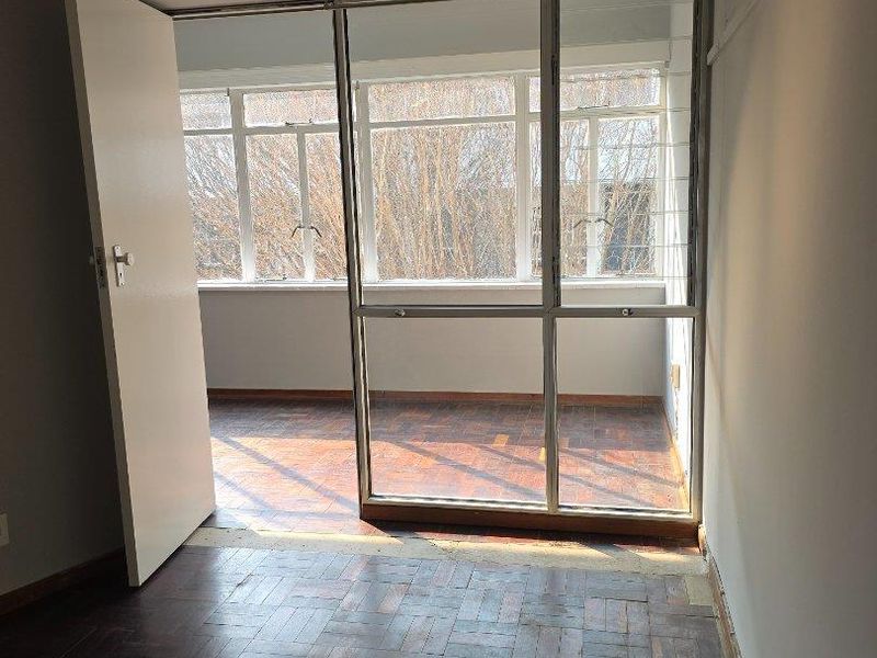 An enclosed balcony or sunroom featuring original parquet flooring and large aluminium-framed windows, offering a bright, versatile space in need of cosmetic refurbishment.