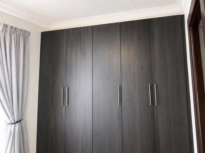 A clean bedroom featuring modern dark wood built-in cupboards, neutral tiled flooring, and classic ceiling cornice detailing.