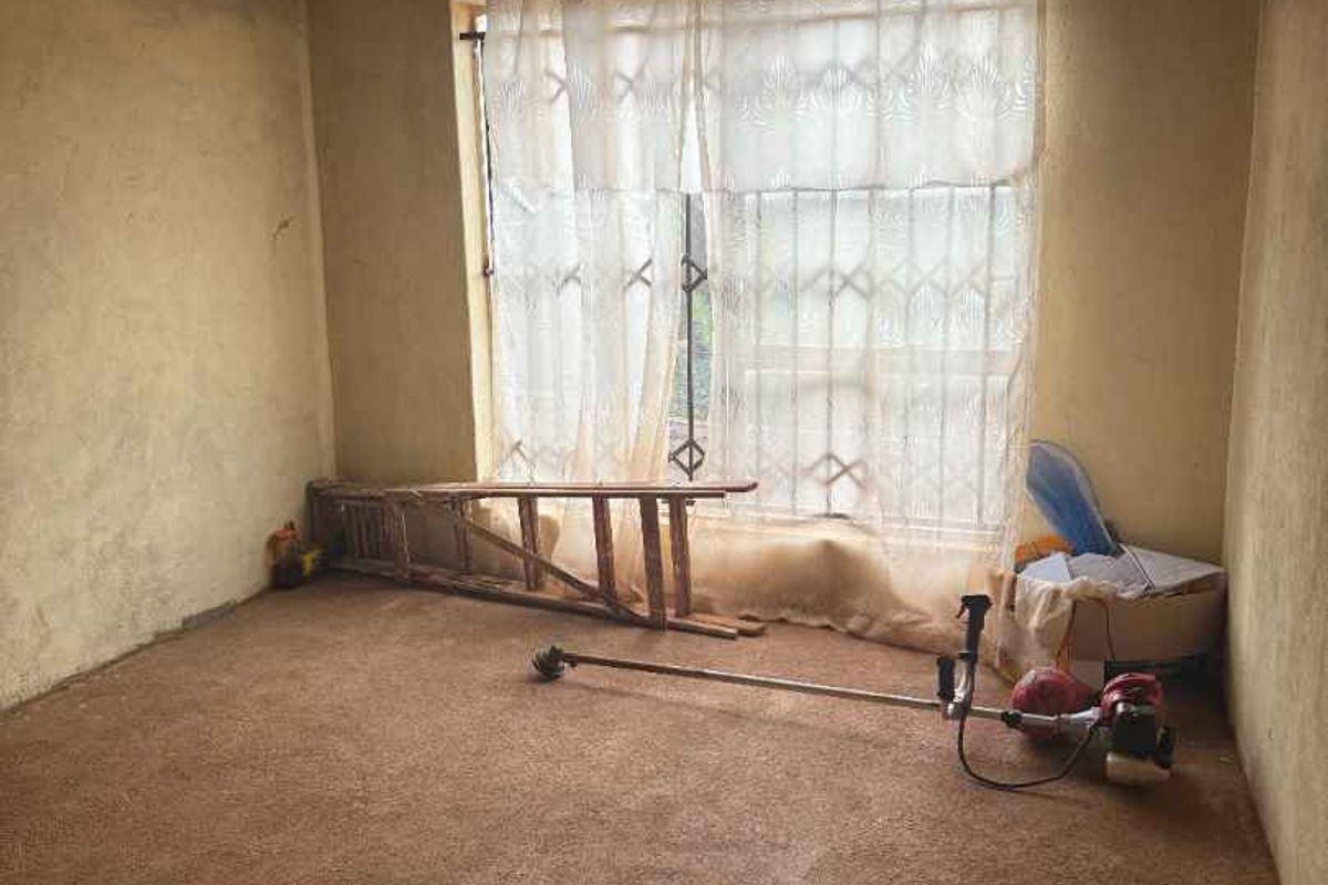 A bedroom space in need of renovation, featuring exposed ceiling rafters, unfinished wall surfaces, and a large window with security bars.