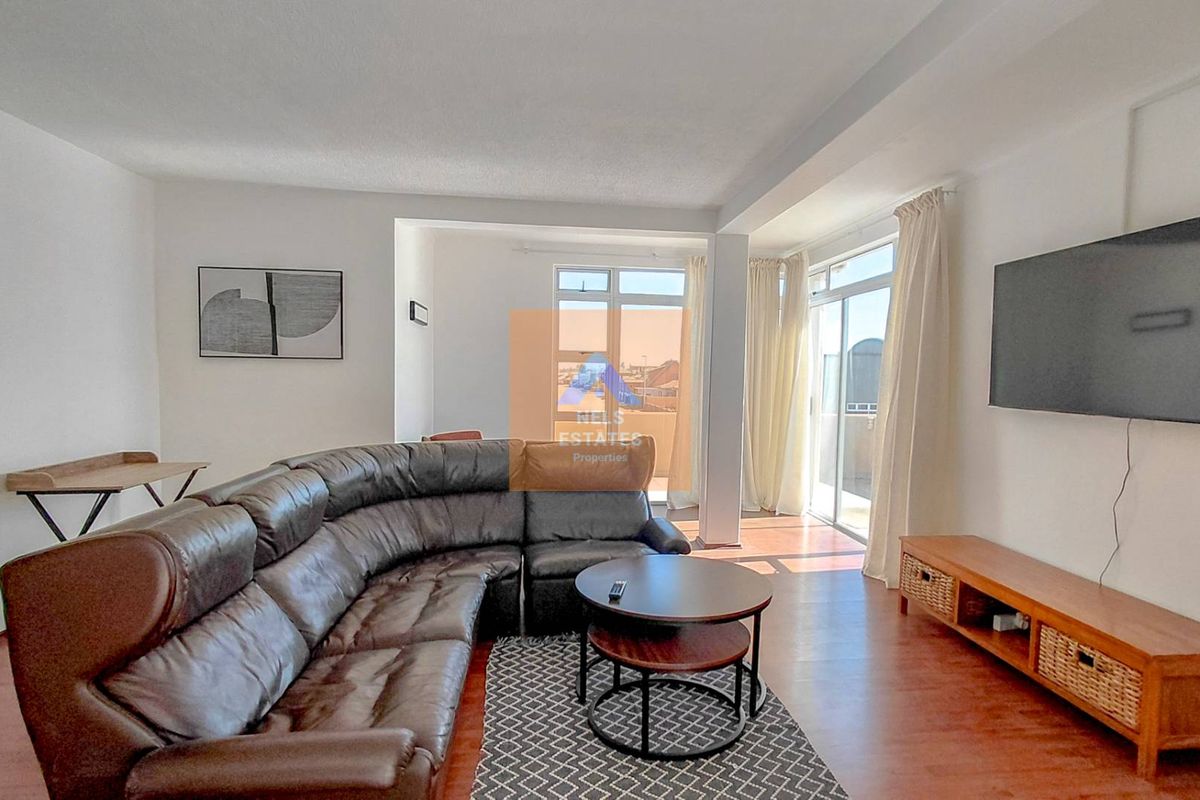 A bright and spacious living room featuring laminate flooring, large sliding glass doors leading to a balcony, and a wall-mounted air conditioning unit.
