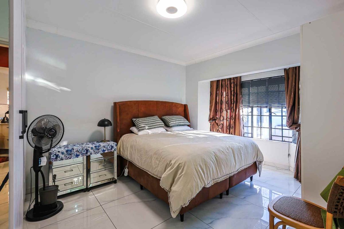 A well-lit bedroom featuring polished floor tiles, a large window with security bars, and a central ceiling light fixture.