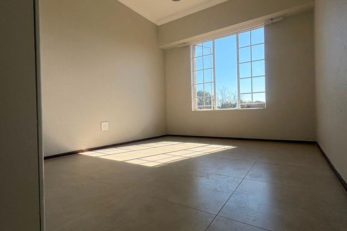 A bright, empty bedroom featuring neutral tiled flooring, a modern pendant light fixture, and a large grid-pattern window allowing for ample natural light.
