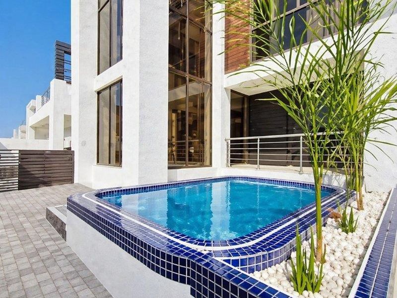 Modern multi-story residence featuring a sleek tiled plunge pool, stone-accented landscaping, and expansive glass windows overlooking a paved patio area.