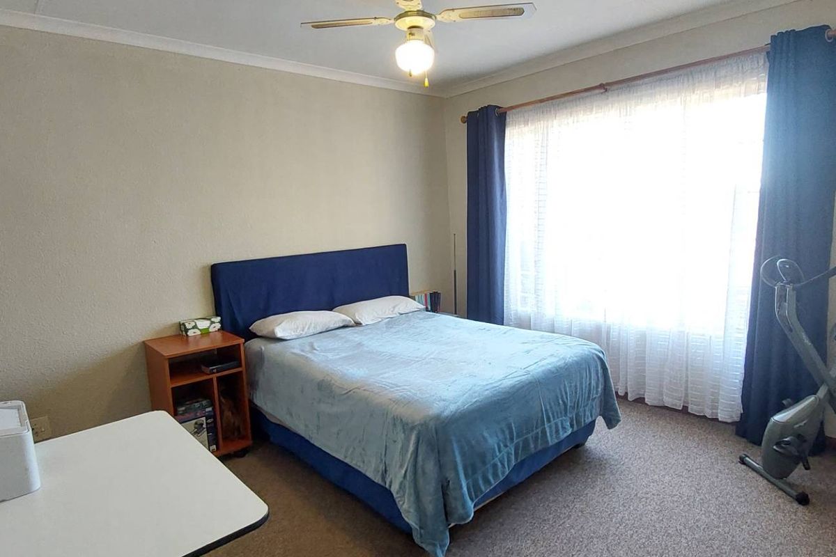 A standard bedroom featuring carpeted flooring, a ceiling fan, and a large window with curtain tracks.