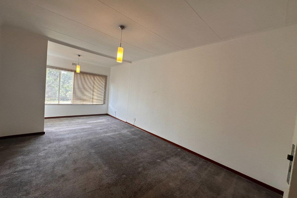 A spacious, empty living area featuring neutral walls, carpeted flooring, and natural light provided by large windows with fitted venetian blinds.