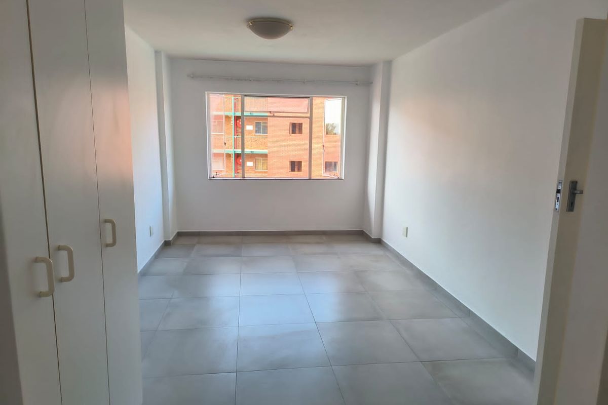 A bright, empty bedroom featuring neutral floor tiles, built-in white cabinetry, and a window providing natural light and a view of the surrounding complex.