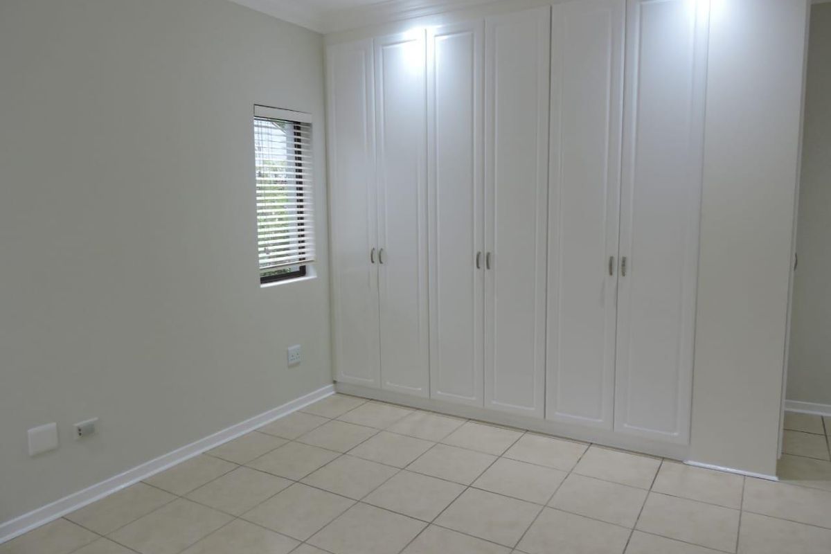 A clean, empty bedroom featuring neutral tiled flooring, built-in white cabinetry, and a window with fitted blinds.
