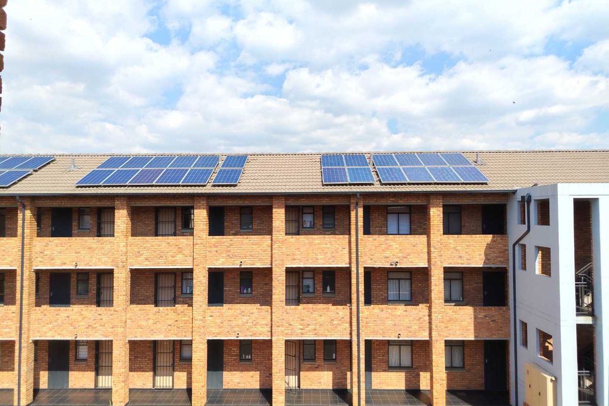 Exterior view of a multi-story apartment building featuring face brick construction, security gates on balconies, and roof-mounted solar panels.