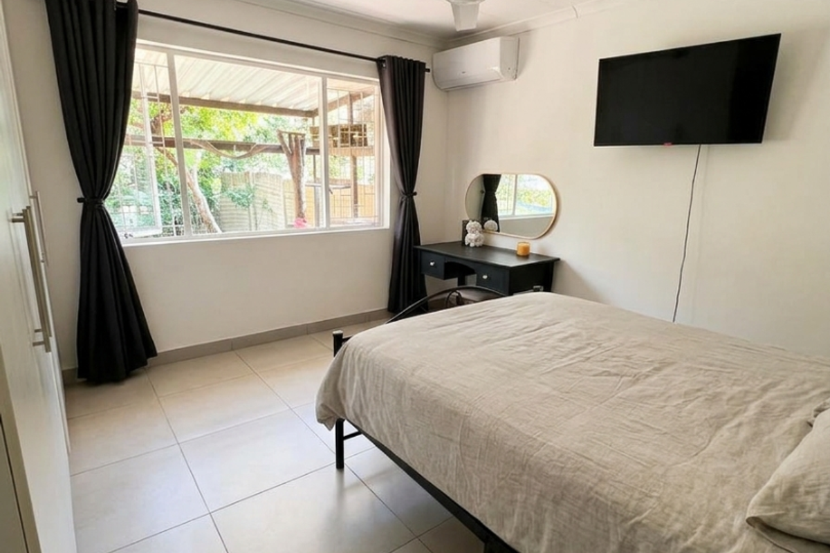 A well-maintained bedroom featuring neutral tiled flooring, a ceiling fan, and a wall-mounted air conditioning unit, offering a clean and functional living space.