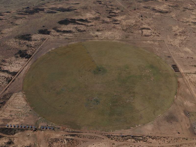 Aerial view of a large circular agricultural field featuring a center pivot irrigation system and a small solar panel array on the property.