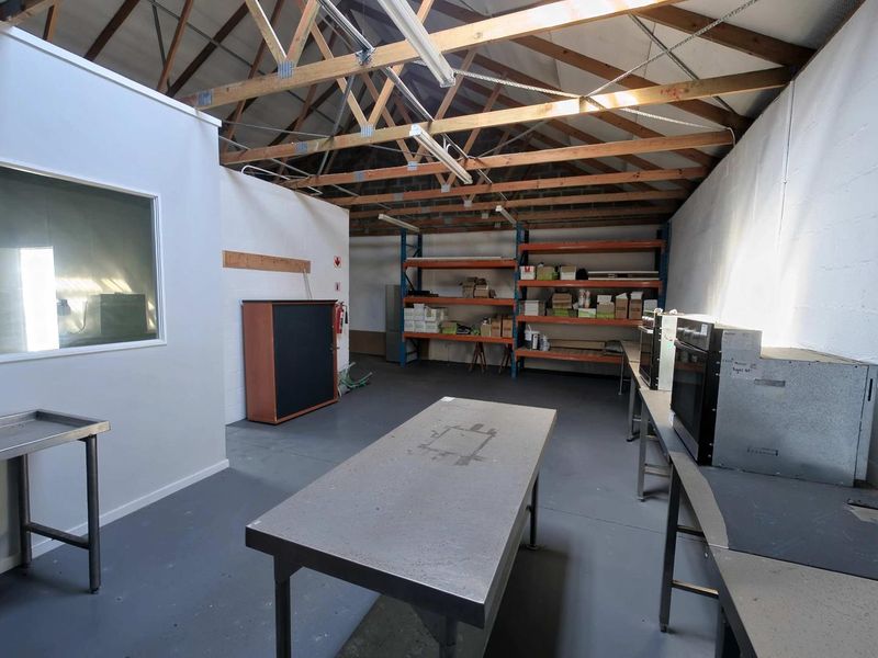 Industrial utility space featuring exposed timber roof trusses, concrete flooring, stainless steel work surfaces, and integrated industrial shelving units.
