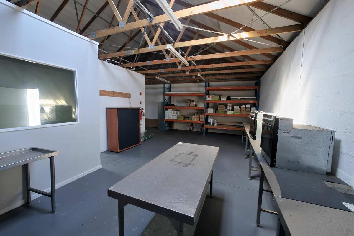 Industrial utility space featuring exposed timber roof trusses, concrete flooring, stainless steel work surfaces, and integrated industrial shelving units.
