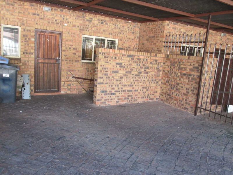 Exterior view of an apartment entrance featuring a brick facade, paved driveway, and a covered carport area with security fencing.
