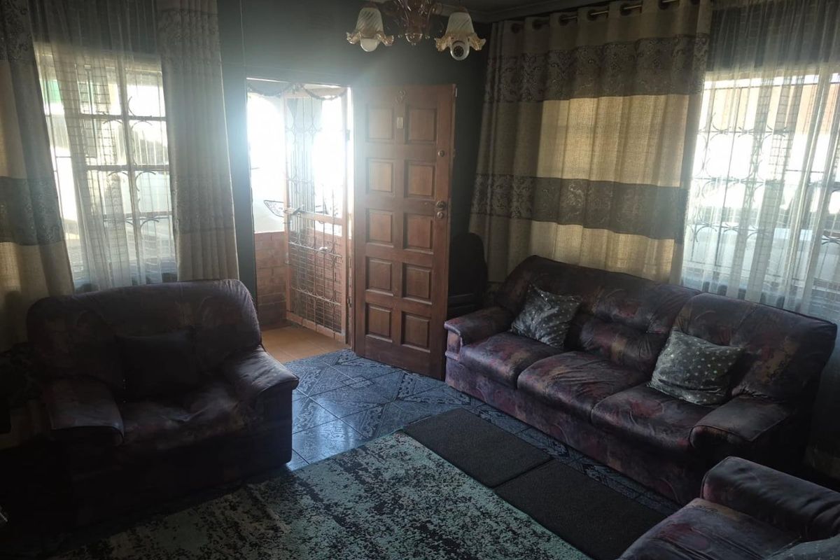 A living room featuring tiled flooring, a timber entrance door with security gate, and natural light from dual windows.