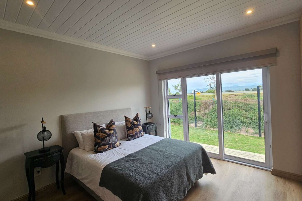 A bright, modern bedroom featuring light wood-look flooring, a tongue-and-groove ceiling, built-in cabinetry, and large sliding glass doors opening to a garden view.