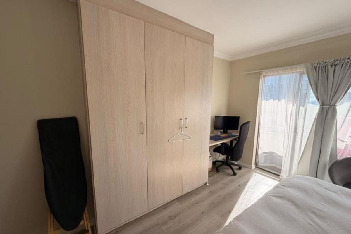 A well-maintained bedroom featuring light wood-tone built-in wardrobes, modern laminate flooring, and a sliding glass door providing natural light.