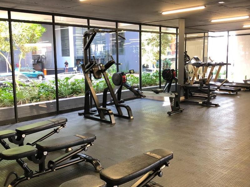 A modern apartment complex gym featuring rubber flooring, floor-to-ceiling windows, and an exposed concrete ceiling with integrated lighting.