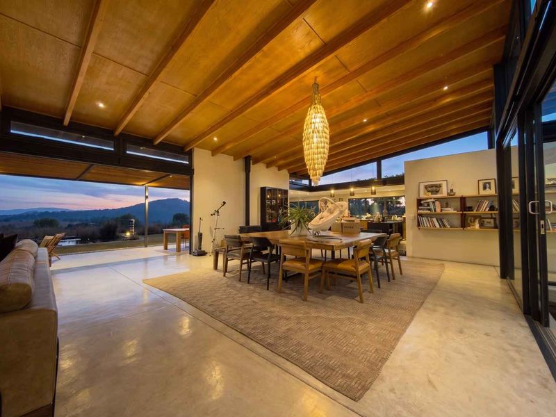 Spacious open-plan living and dining area featuring high exposed timber ceilings, polished concrete floors, and expansive sliding glass doors with scenic mountain views.
