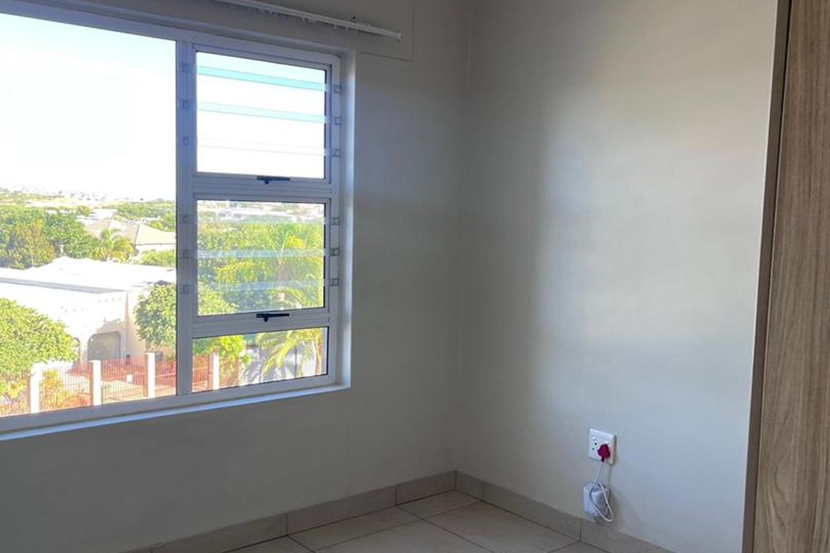 A bright, empty bedroom featuring neutral tiled flooring, modern track lighting, and a large window offering an elevated view of the surrounding neighborhood.