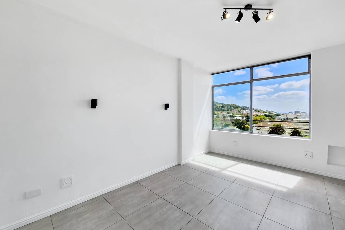A bright, modern, and empty living area featuring large format floor tiles, track lighting, and a large window offering expansive neighborhood views.