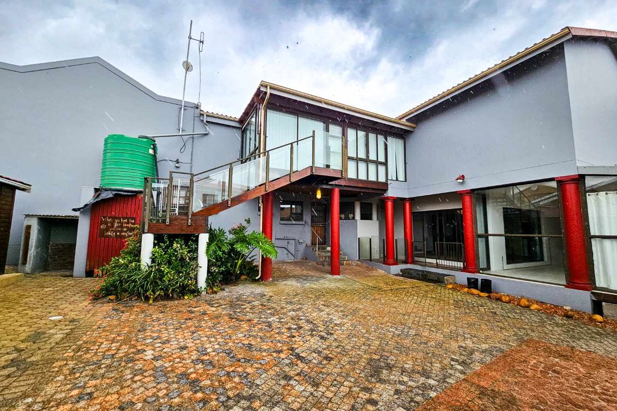 Rear exterior view of the property featuring a paved courtyard, elevated balcony with glass balustrade, and red structural columns.
