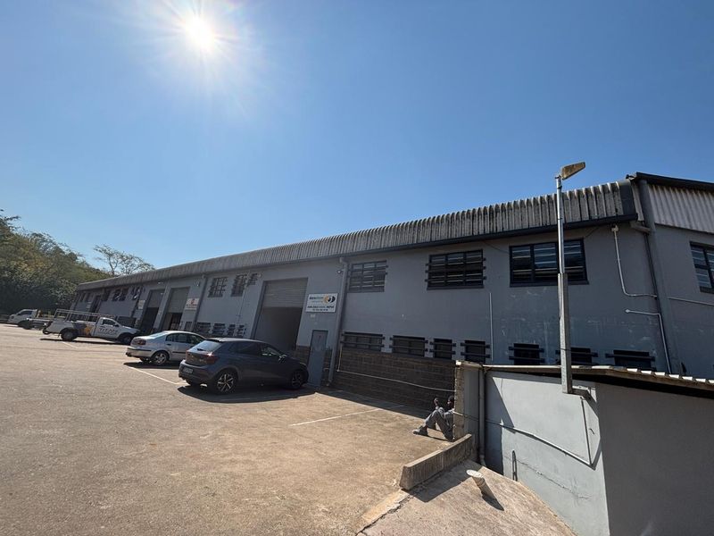 Front elevation of an industrial warehouse facility featuring roller shutter access, metal roofing, and an adjacent paved parking area.