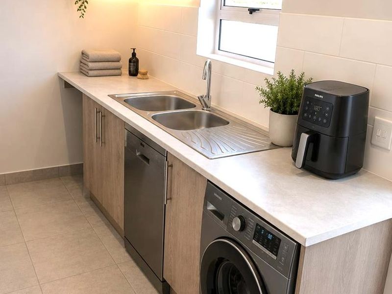 Modern laundry room featuring light wood-look cabinetry, stone-effect countertops, a stainless steel double sink, and integrated under-shelf lighting.