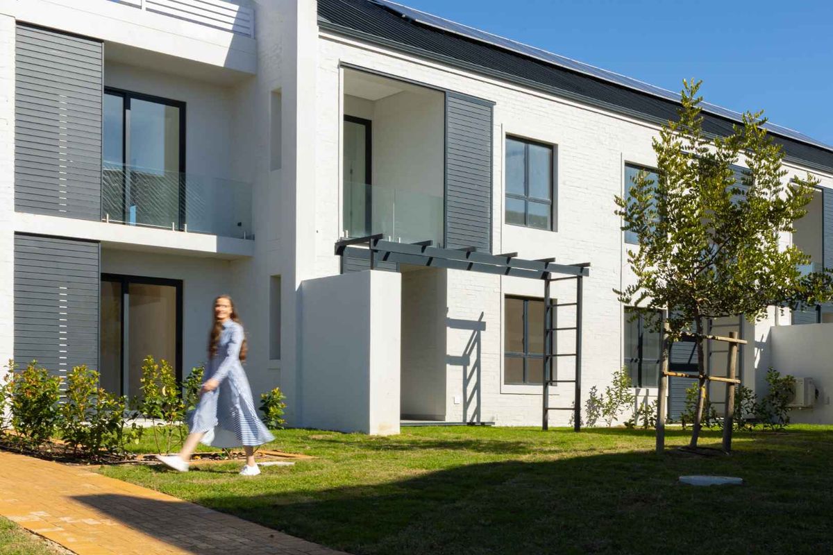 Rear view of a modern apartment building featuring white brick facade, glass balcony railings, and a metal pergola over a paved patio area.