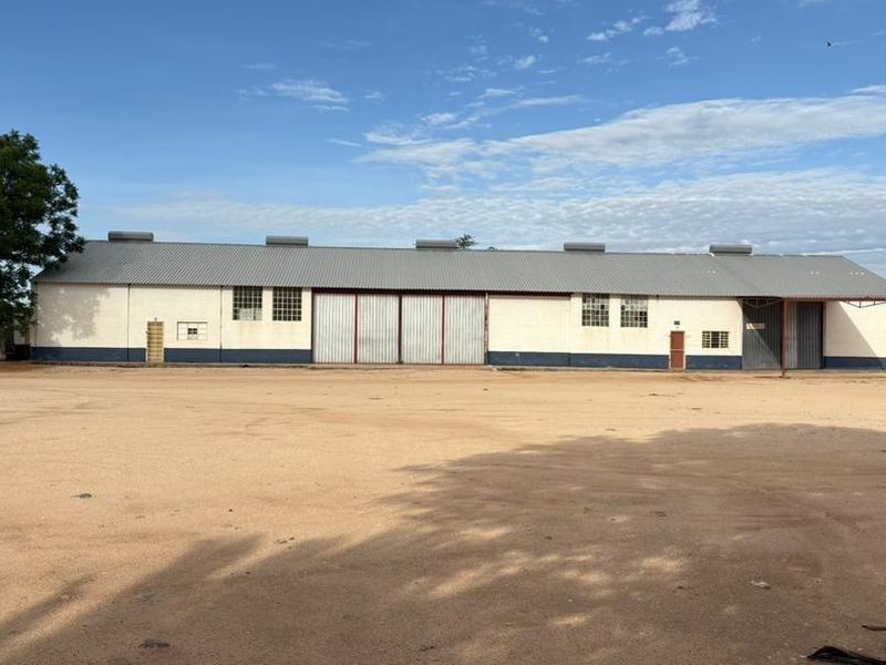 A wide-angle view of a commercial warehouse building featuring a large metal roof, industrial sliding doors, and a spacious unpaved yard area.