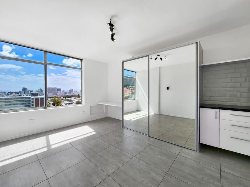 Bright and modern bedroom featuring large windows with city views, tiled flooring, a built-in desk, and a spacious mirrored sliding wardrobe.